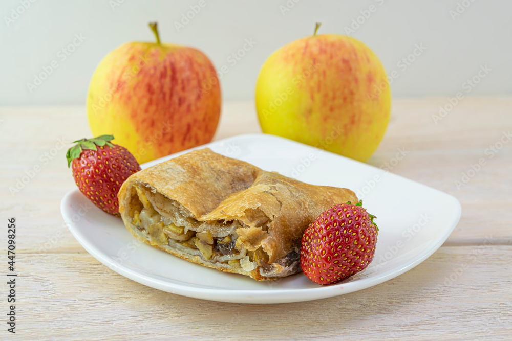 A piece of apple strudel with cinnamon in a white plate with strawberries close-up A piece of apple strudel with cinnamon in a white plate with strawberries close-up