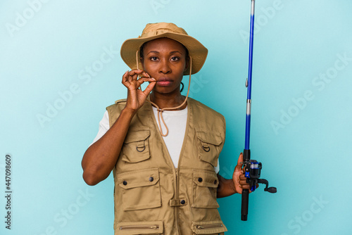 Young african american fisherwoman holding rod isolated on blue background with fingers on lips keeping a secret.