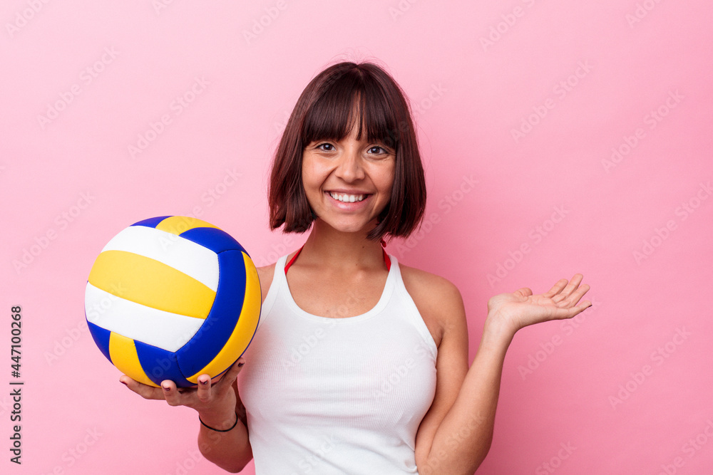 Young mixed race woman playing volleyball isolated on pink background showing a copy space on a palm and holding another hand on waist. Young mixed race woman playing volleyball isolated on pink background showing a copy space on a palm and holding another hand on waist.