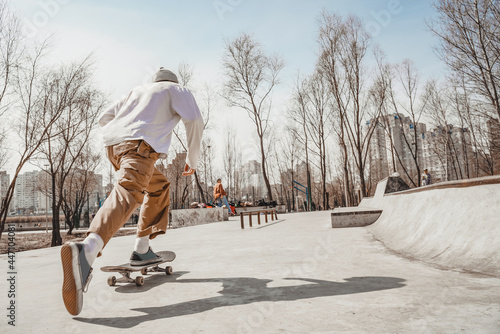 Skater guy rides a skateboard very quickly, pushing off the ground with his right foot, goes to his skater friends. The sun reflects the shadow of a skater on the gray surface of a skatepark.