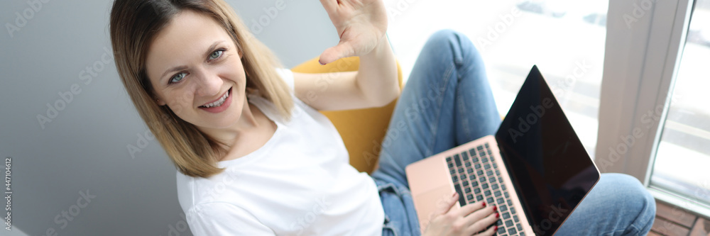 Fototapeta premium Young woman with laptop sitting in armchair and waving hand