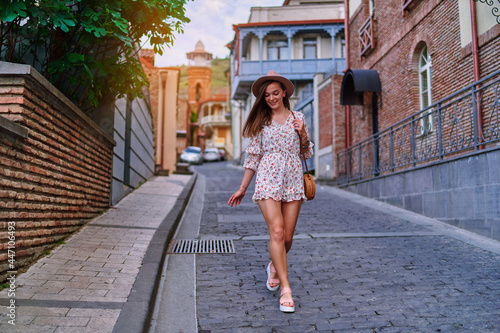 Young happy smiling cute beautiful brunette traveler girl with long legs wearing hat, round straw bag and short jumpsuit walking alone around the city at sunny summer day © Goffkein