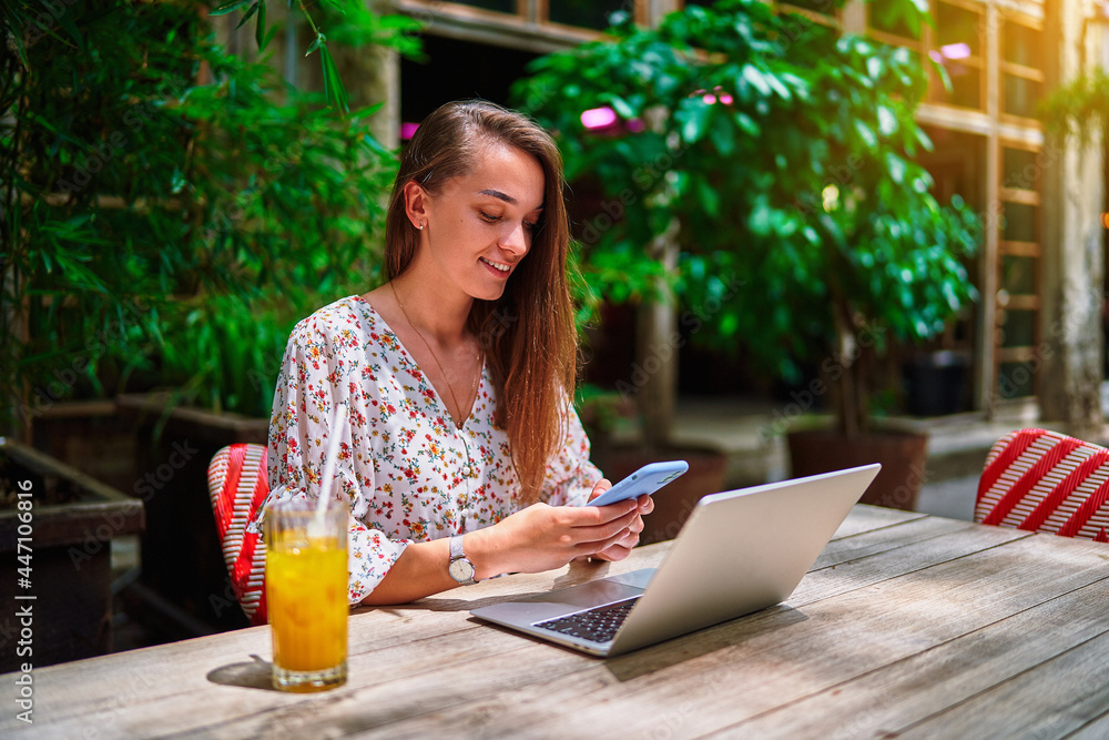 Fototapeta premium Happy cheerful smiling beautiful cute joyful young millennial girl using laptop and phone for remote work, chatting, browsing online and social networking at green cafe