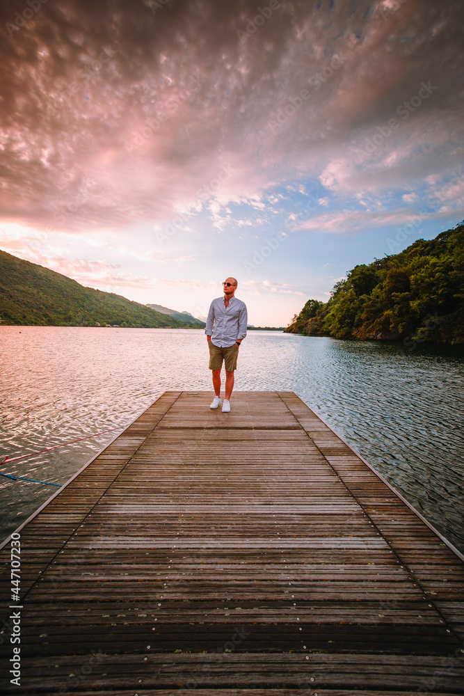 Mergozzo, Verbania / Italy - June 2021: Young man walking on the wooden jetty of Lake Mergozzo at dawn with colorful clouds in the background Mergozzo, Verbania / Italy - June 2021: Young man walking on the wooden jetty of Lake Mergozzo at dawn with colorful clouds in the background