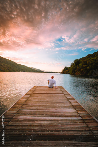 Mergozzo, Verbania / Italy - June 2021: Young man sitting on the wooden jetty of Lake Mergozzo at dawn with colorful clouds in the background