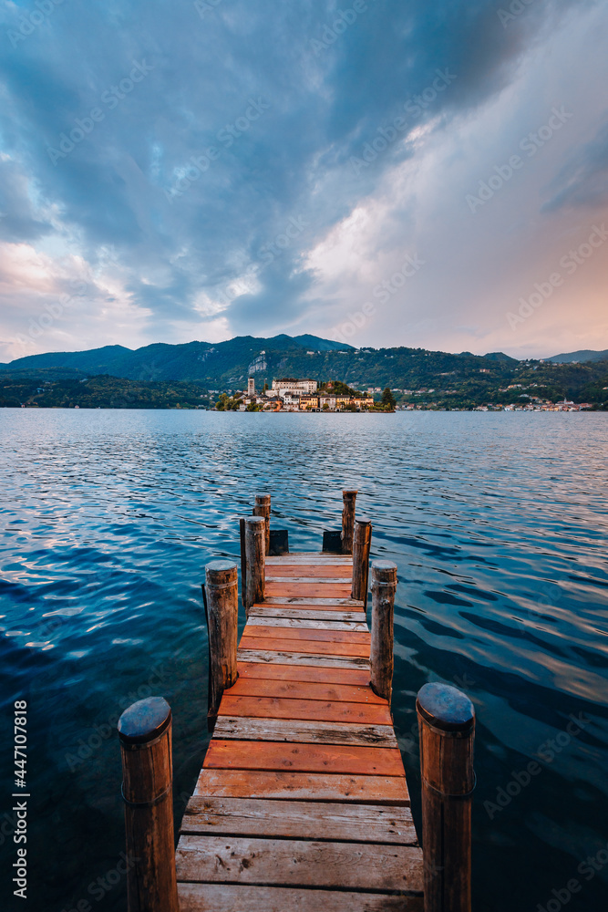 Orta San Giulio / Italy - June 2021: The island of San Giulio with a wooden jetty in the foreground at sunset
