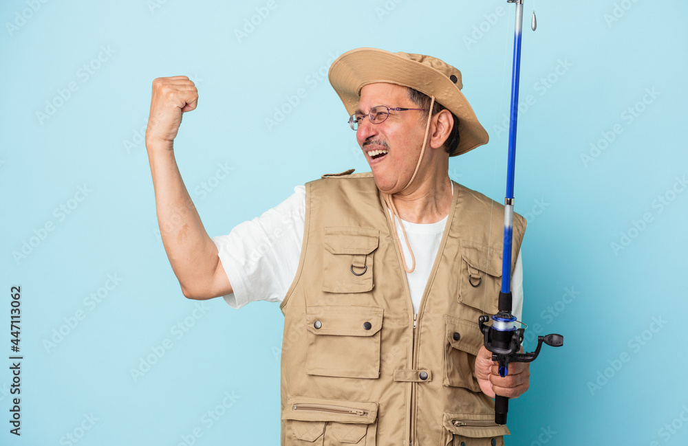 Senior indian fisherman holding rod isolated on blue background raising fist after a victory, winner concept. Senior indian fisherman holding rod isolated on blue background raising fist after a victory, winner concept.