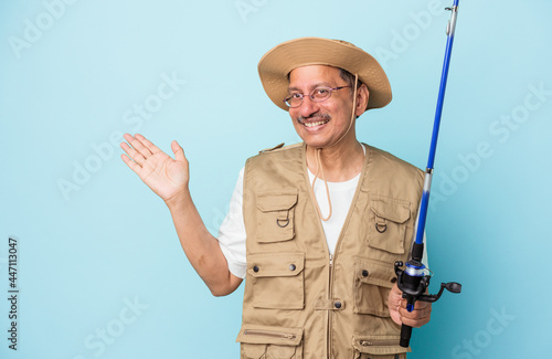 Senior indian fisherman holding rod isolated on blue background showing a copy space on a palm and holding another hand on waist.
