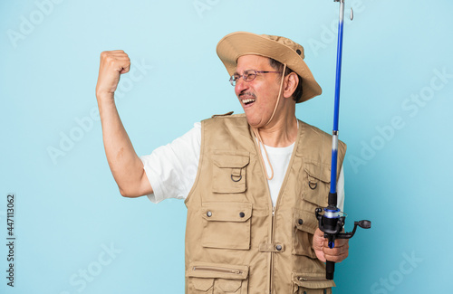 Senior indian fisherman holding rod isolated on blue background raising fist after a victory, winner concept.