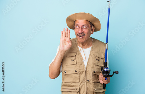 Senior indian fisherman holding rod isolated on blue background is saying a secret hot braking news and looking aside