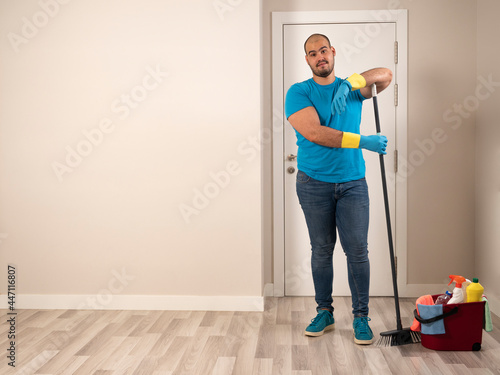 Young man cleaning with a sweeping brush and mop and bucket of water and cleaning supplies at home wearing blue gloves and blue shirt