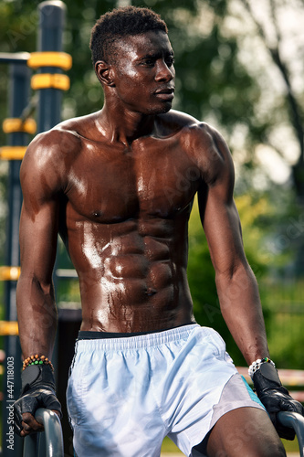 Muscular young man exercising at the sports field. African man looking to the side while doing horizontal bar exercises. Shirtless male model exercising outdoors.