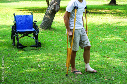 Ankle injury patients wrapped with a bandage ankle brace and use crutches to support walking on the lawn, Which has a wheelchair behind. Healthcare and medical concept.