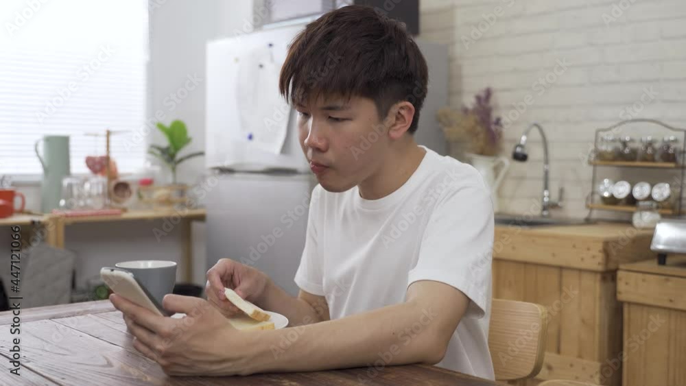 young Asian man holding a mobile phone at the dining table is reading online news with concentration while eating breakfast in the morning. young Asian man holding a mobile phone at the dining table is reading online news with concentration while eating breakfast in the morning.
