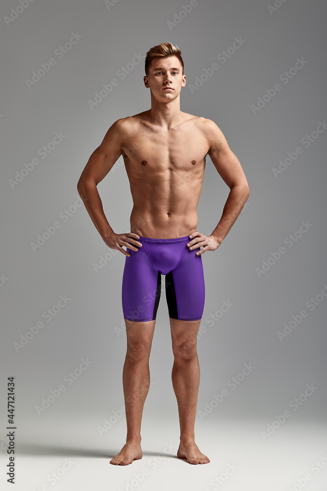 Young attractive swimmer in excellent physical shape in purple swimming shorts, on a gray background, copy space Young attractive swimmer in excellent physical shape in purple swimming shorts, on a gray background, copy space