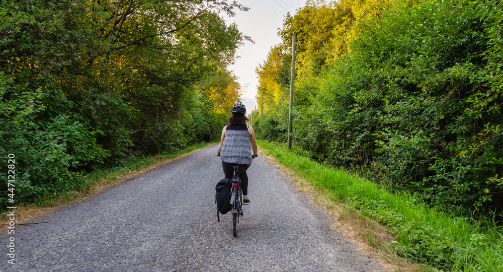 Fototapeta premium Adventurous White Cacasusian Woman riding a bicycle on a road. Sunny Summer Sunset Art Render. Barnston Island, Vancouver, British Columbia, Canada. Adventure Journey Concept