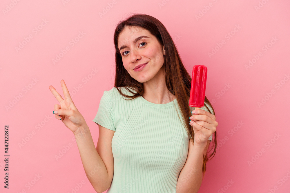 Young caucasian woman holding an ice cream isolated on blue background joyful and carefree showing a peace symbol with fingers. Young caucasian woman holding an ice cream isolated on blue background joyful and carefree showing a peace symbol with fingers.