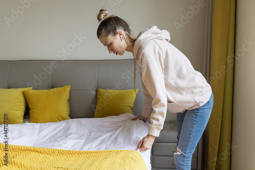 Young caucasian woman using mobile phone and wireless headphones, resting, sitting on bed at home and listening podcast or clubhouse - voice-only social media app, drop-in audio chat