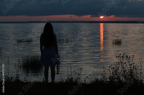 Silhouette of a person on the lake at sunset with a musical instrument a trumpet looking at the landscape the hot sun and the reflection of a red illuminated path on the water in summer at sunset