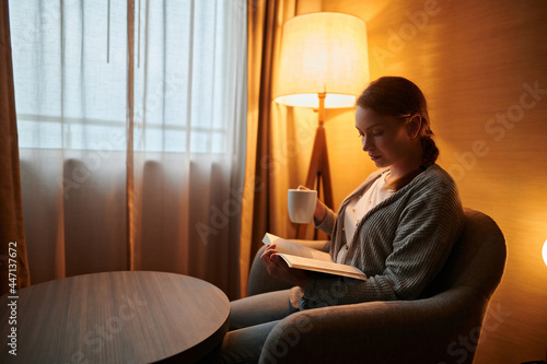 Cozy evening at home.A young woman is sitting in a chair and reading a book. She has a cup of tea in her hand. In the corner of the room, a floor lamp burns with a soft yellow light.