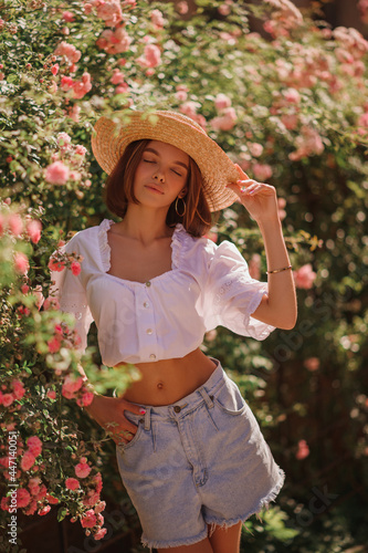 Fashionable dreamful woman with closed eyes wearing trendy summer outfit with straw hat, stylish white blouse, denim shorts, posing in street, near blooming roses