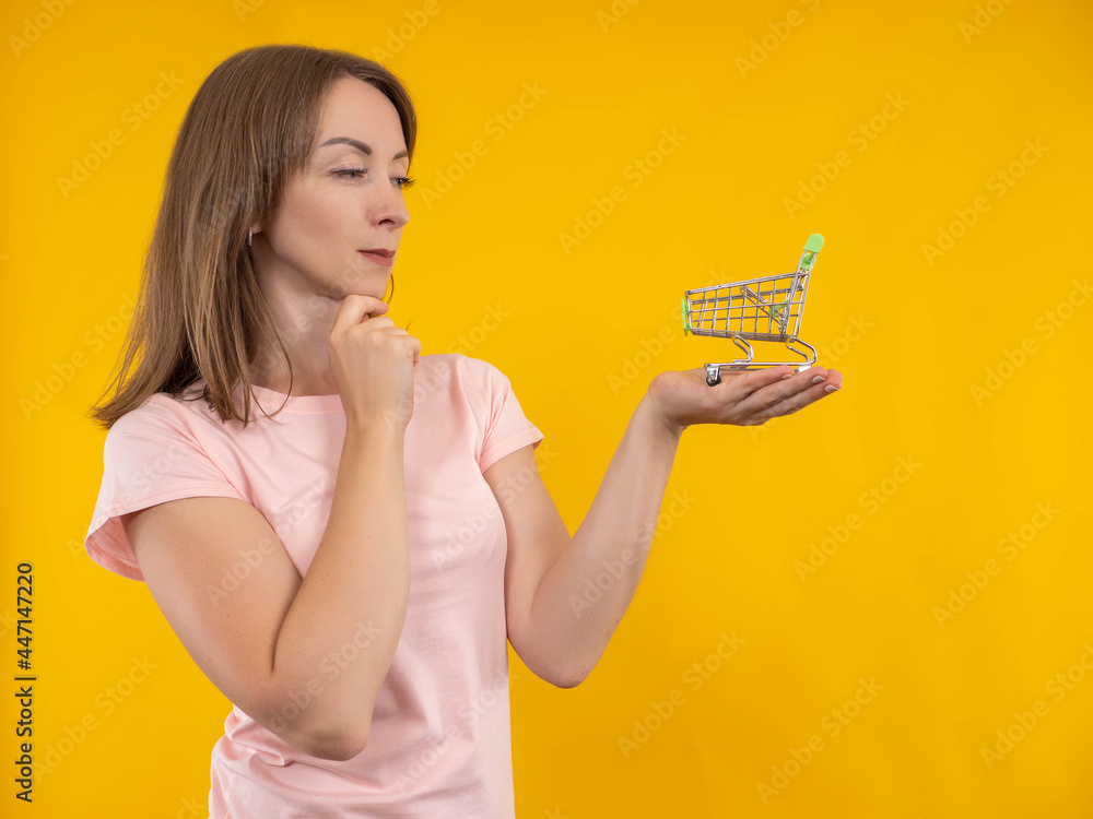 Woman thought about shopping. Girl is thinking about shopping. Shopper girl on yellow background. Concept - she chooses where to make purchase. Woman shopper with trolley from supermarket. Woman thought about shopping. Girl is thinking about shopping. Shopper girl on yellow background. Concept - she chooses where to make purchase. Woman shopper with trolley from supermarket.