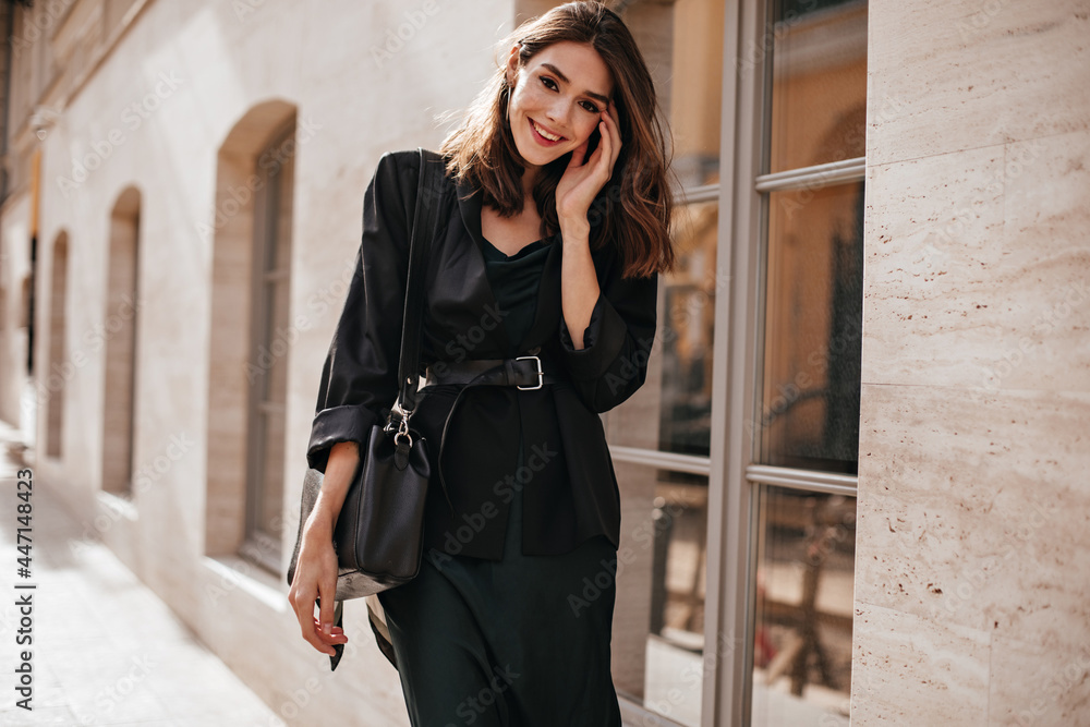 Cute slim girl with brown wavy hairstyle, bright makeup, dark outfit of long dress, oversized jacket, belt on waist and trendy bag, standing on daylight street in city and smiling
