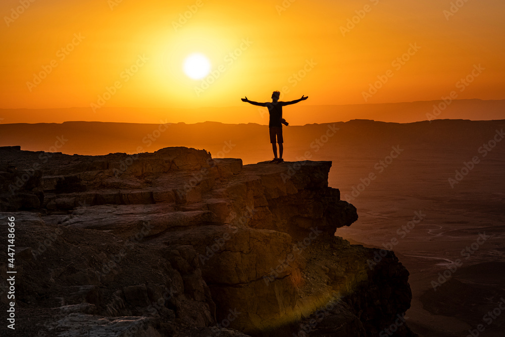 Man standing on a cliff with his arms open and looking at the sun rise over the Negev Desert in Israel - obrazy, fototapety, plakaty 