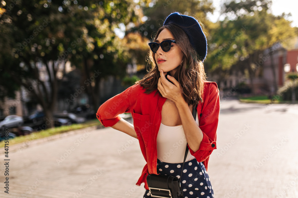 Profile portrait of young lady with brunette wavy hair, beret and sunglasses, wearing white top and red shirt, posing on sunny street against green trees background
