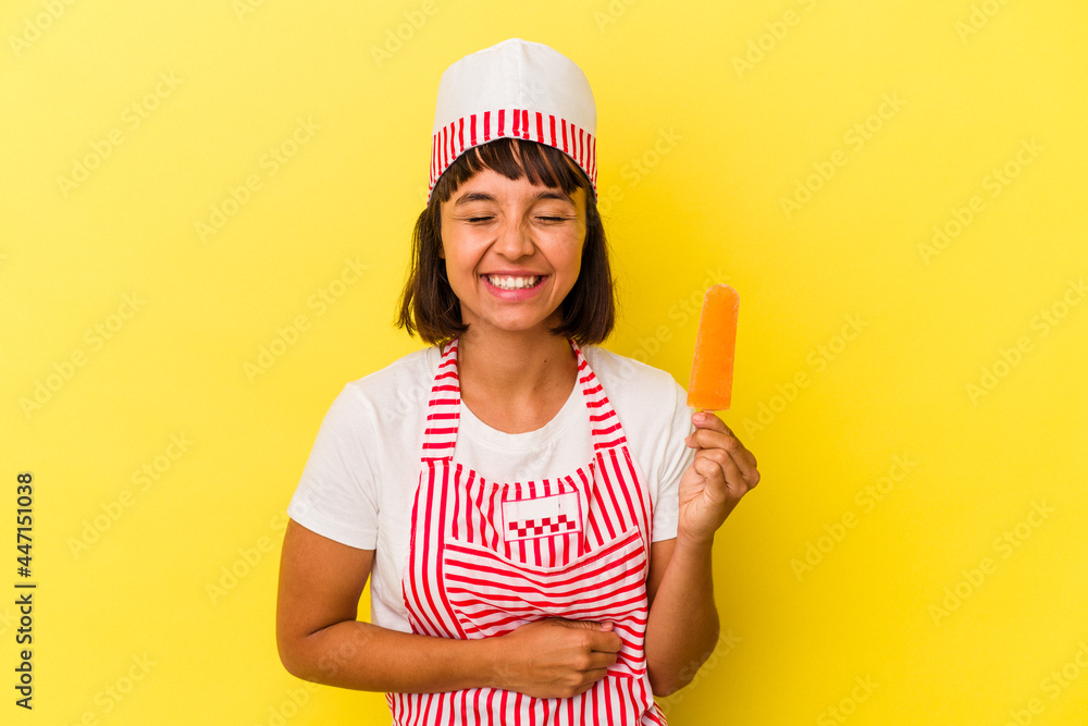 Young mixed race ice cream maker woman holding an ice cream isolated on yellow background laughing and having fun. Young mixed race ice cream maker woman holding an ice cream isolated on yellow background laughing and having fun.