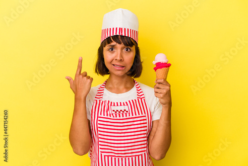 Young mixed race ice cream maker woman holding an ice cream isolated on yellow background showing a disappointment gesture with forefinger. © Asier