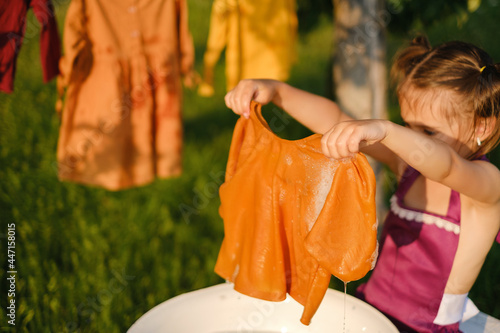 The girl is holding a wet soapy shirt in foam in her hands. The child washes clothes outdoors and hangs them to dry on a clothesline. Hand washing of colored underwear