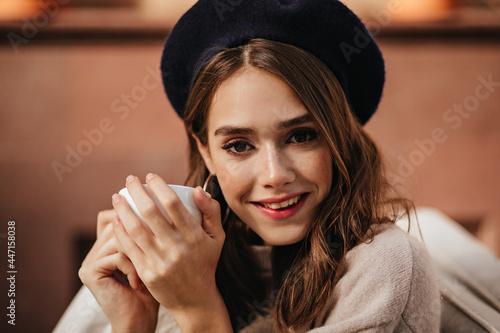Pretty young lady with brunette wavy hair and dark eyes, trendy makeup, in beret and pullover sitting outdoors with cup of tea and smiling