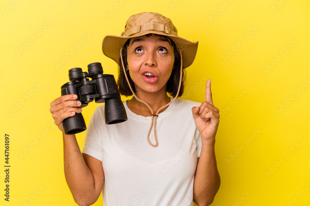Young mixed race explorer woman holding binoculars isolated on yellow background pointing upside with opened mouth. Young mixed race explorer woman holding binoculars isolated on yellow background pointing upside with opened mouth.
