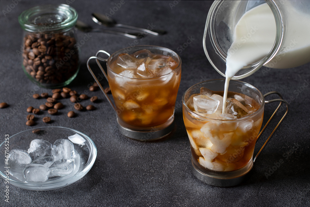 Two cups of cold coffee with ice cubes, into which milk is being poured from a jug. Dark background. Close-up. Two cups of cold coffee with ice cubes, into which milk is being poured from a jug. Dark background. Close-up.