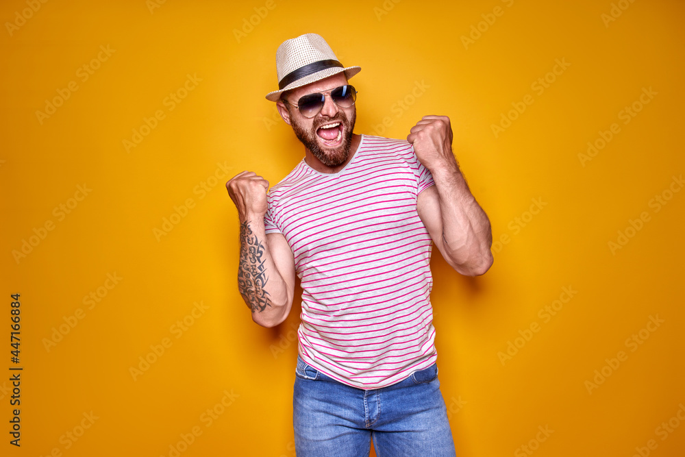 Image of excited bearded tattooed man in sunglasses and hat making winner gesture isolated yellow background.