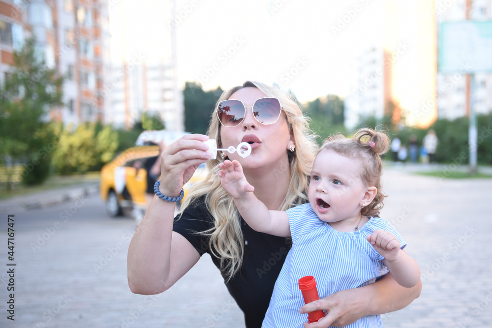 A young blonde mom in sunglasses teaches her little daughter to blow and blow soap bubbles
