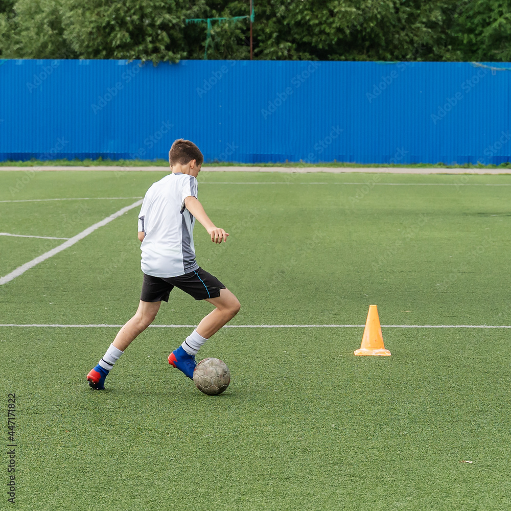 The boy loves to play football, trains with the ball on the artificial turf and scores a goal. The boy plays football