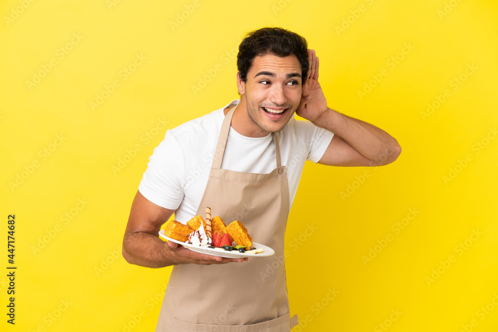 Restaurant waiter holding waffles over isolated yellow background listening to something by putting hand on the ear