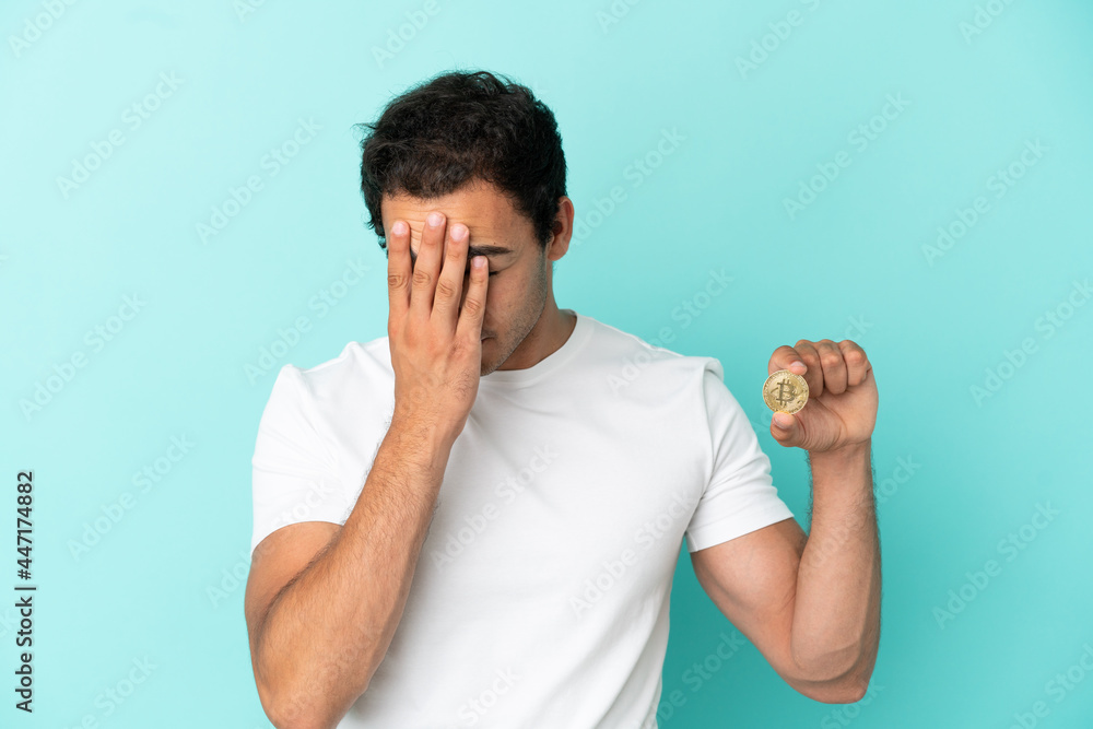 Young man holding a Bitcoin over isolated blue background with tired and sick expression Young man holding a Bitcoin over isolated blue background with tired and sick expression