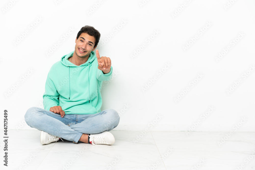 Caucasian handsome man sitting on the floor showing and lifting a finger Caucasian handsome man sitting on the floor showing and lifting a finger