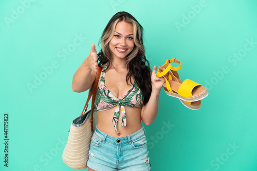 Teenager Russian girl holding summer sandals and beach bag isolated on green background shaking hands for closing a good deal