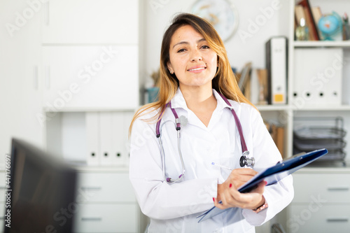 Portrait of smiling hispanic female doctor writing notes on clipboard in clinic office