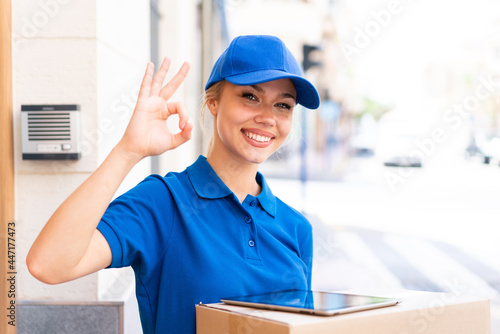 Young delivery woman at outdoors holding boxes and a tablet and doing OK sign