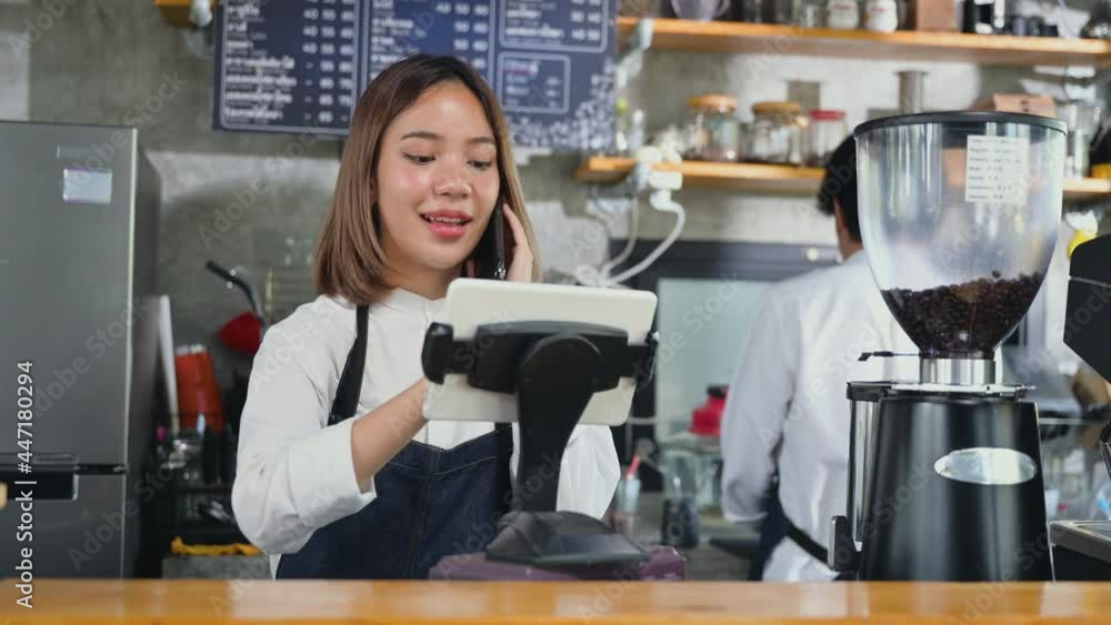 Owner coffee shop and online order concept. Asian young woman barista in apron talking on mobile phone and using tablet to customer in coffee shop. Small business cafe restaurant retail food and drink Owner coffee shop and online order concept. Asian young woman barista in apron talking on mobile phone and using tablet to customer in coffee shop. Small business cafe restaurant retail food and drink