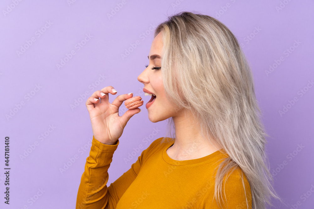 Teenager girl over isolated purple background holding colorful French macarons and eating it
