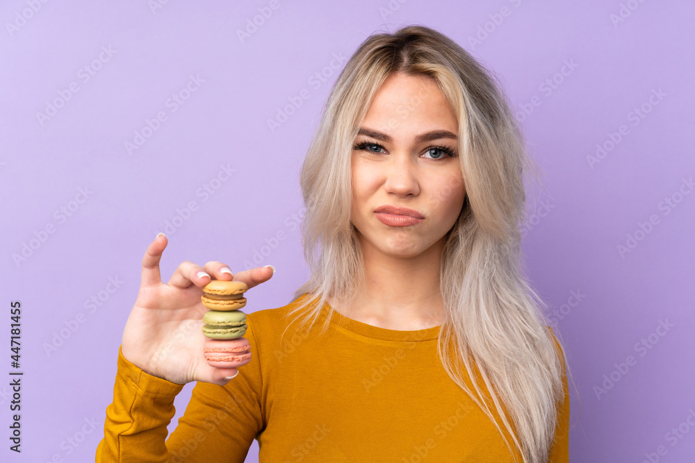 Teenager girl over isolated purple background holding colorful French macarons and with sad expression Teenager girl over isolated purple background holding colorful French macarons and with sad expression