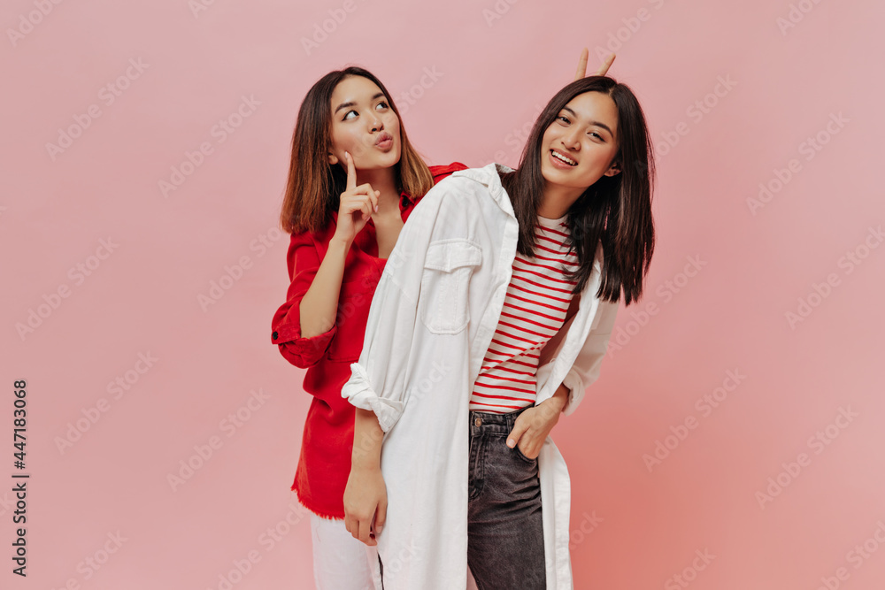 Cute Asian women in oversized shirts make funny faces on isolated background. Charming girl in red blouse looks thoughtful and gives bunny ears to her friend. Brunette woman on pink backdrop.