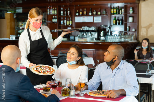 Polite waitress in protective mask bringing ordered pizza to friends visited restaurant for lunch © JackF