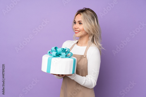 Young pastry chef holding a big cake over isolated purple background looking to the side